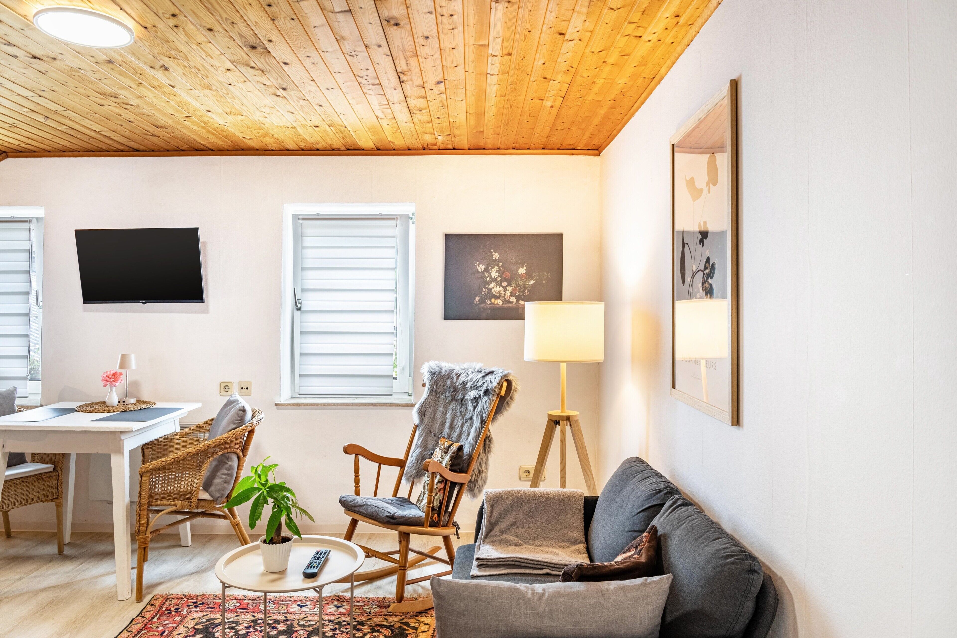 En hyggelig stue med træloft, fjernsyn, sofa, lænestol, gulvlampe og et billede på væggen.