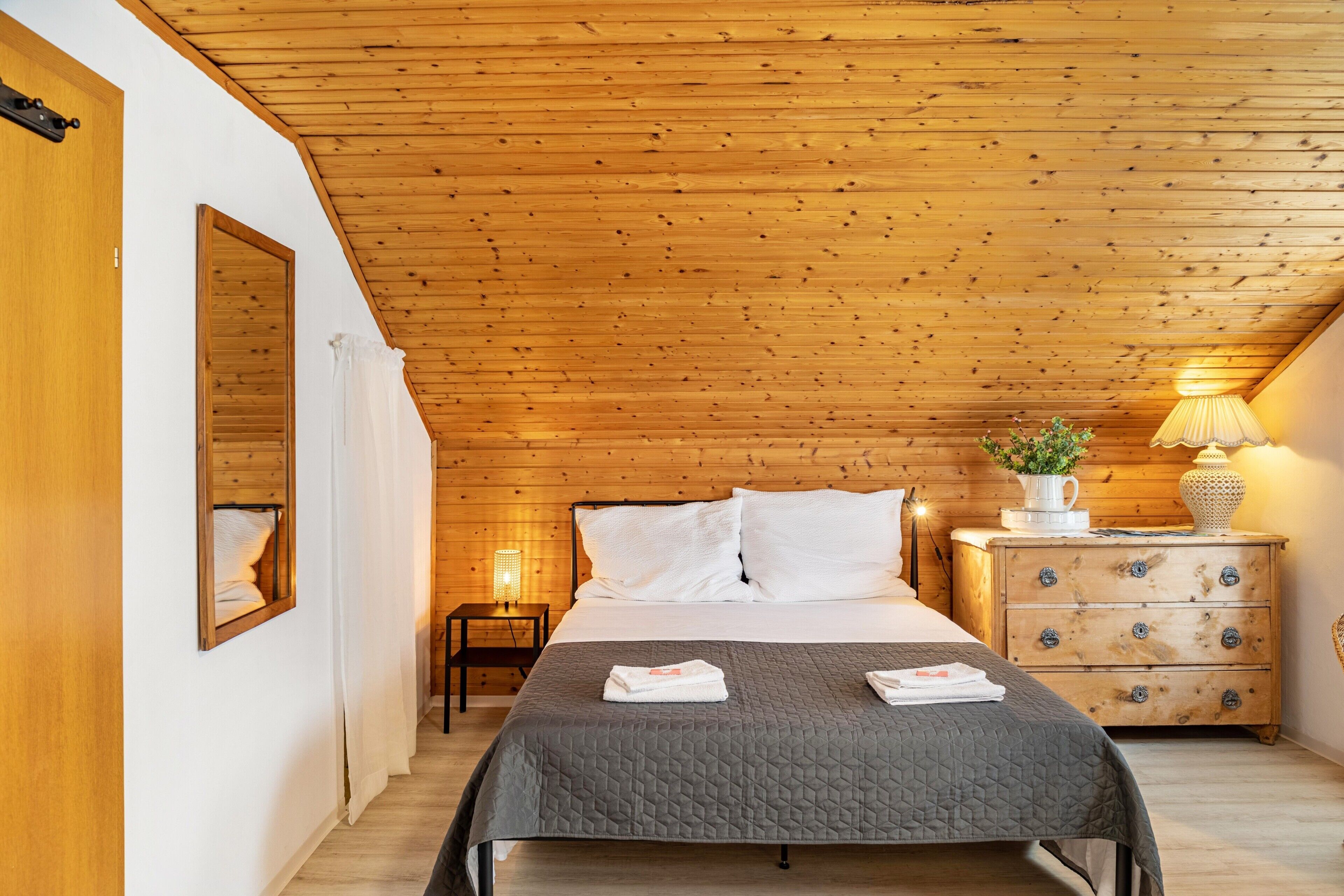 A bedroom with a wooden ceiling, a bed with white bedding, a nightstand with a lamp, and a dresser with drawers.