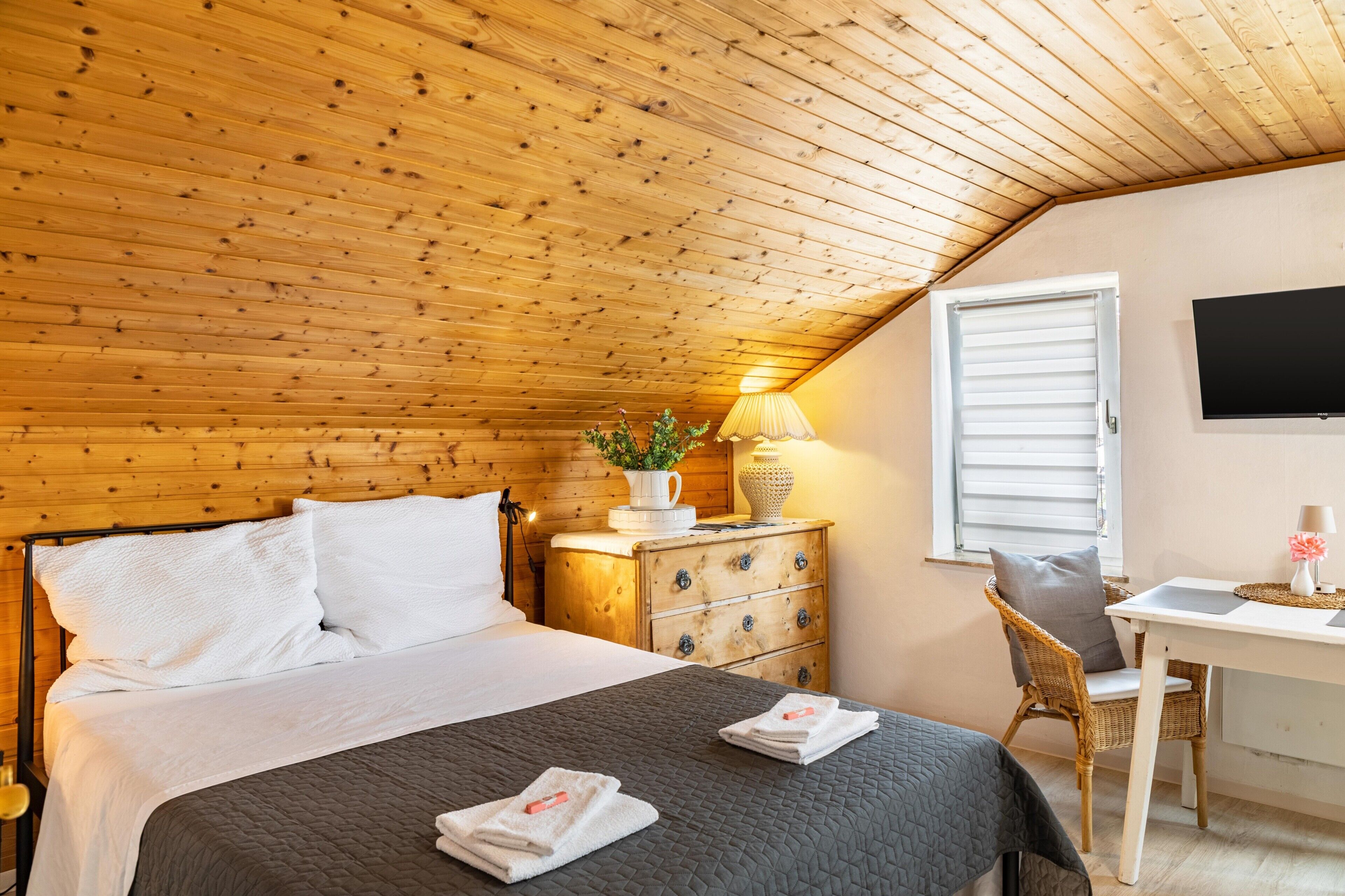 Un dormitorio con techo de madera, una cama con ropa blanca, una mesita de noche con lámpara, una pequeña mesa con una silla y un televisor montado en la pared.