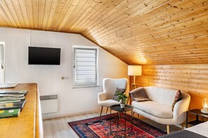 Tunnelmallinen huone, jossa on kalteva katto, seinälle asennettu taulutelevisio, sälekaihtimilla varustettu ikkuna, pehmeä nojatuoli, pieni pöytä, jossa on kasvi, ja lamppu.