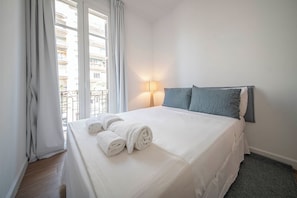 A hotel room with a bed, white linens, a lamp, and a view of buildings through the window. - 3 Bedroom Apartment Uptown Barcelona (Barcelona)