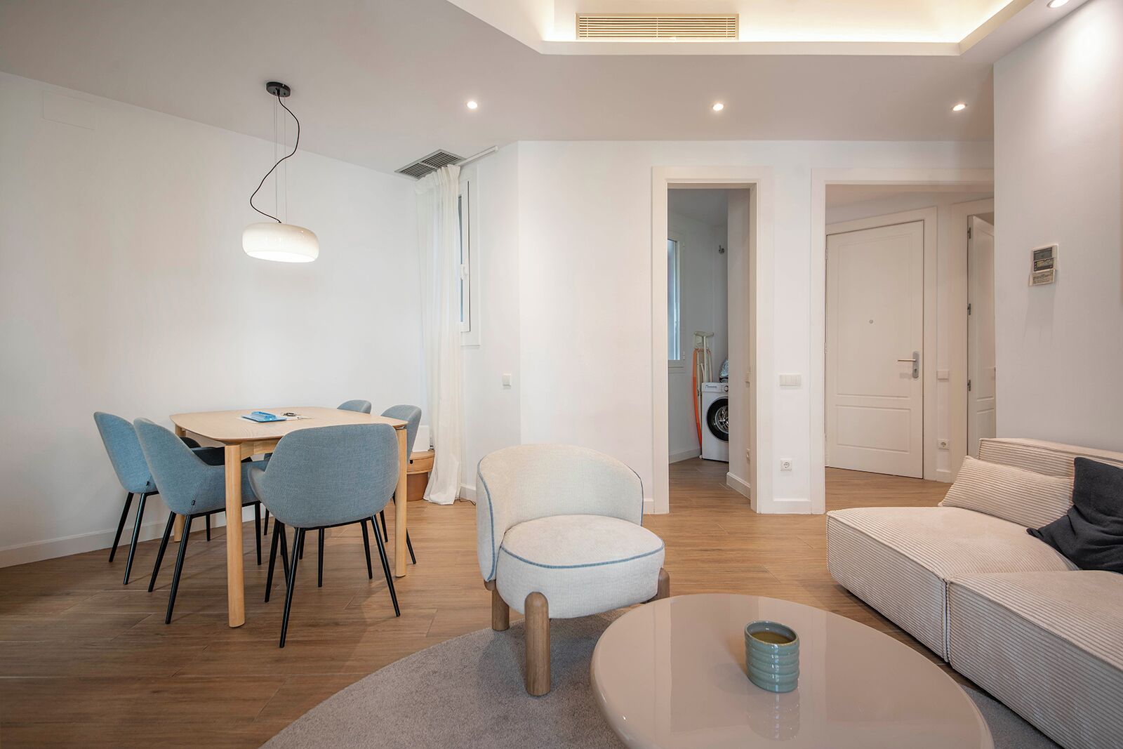 A modern living room with a dining area, a sofa, a coffee table, and a washing machine in the background.