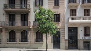Ein mehrstöckiges Gebäude mit einem Baum davor, Balkonen mit Metallgeländern und einer Tür mit einer Hausnummer.