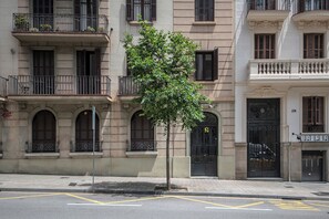 Ein mehrstöckiges Gebäude mit einem Baum davor, Balkonen mit Metallgeländern und einer Tür mit einer Hausnummer.
