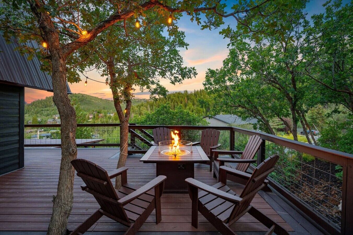 A deck with a fire pit table and chairs, surrounded by trees and overlooking a landscape.