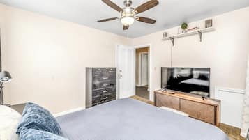 A bedroom with a bed, a ceiling fan, a TV, a dresser, and a door leading to another room.