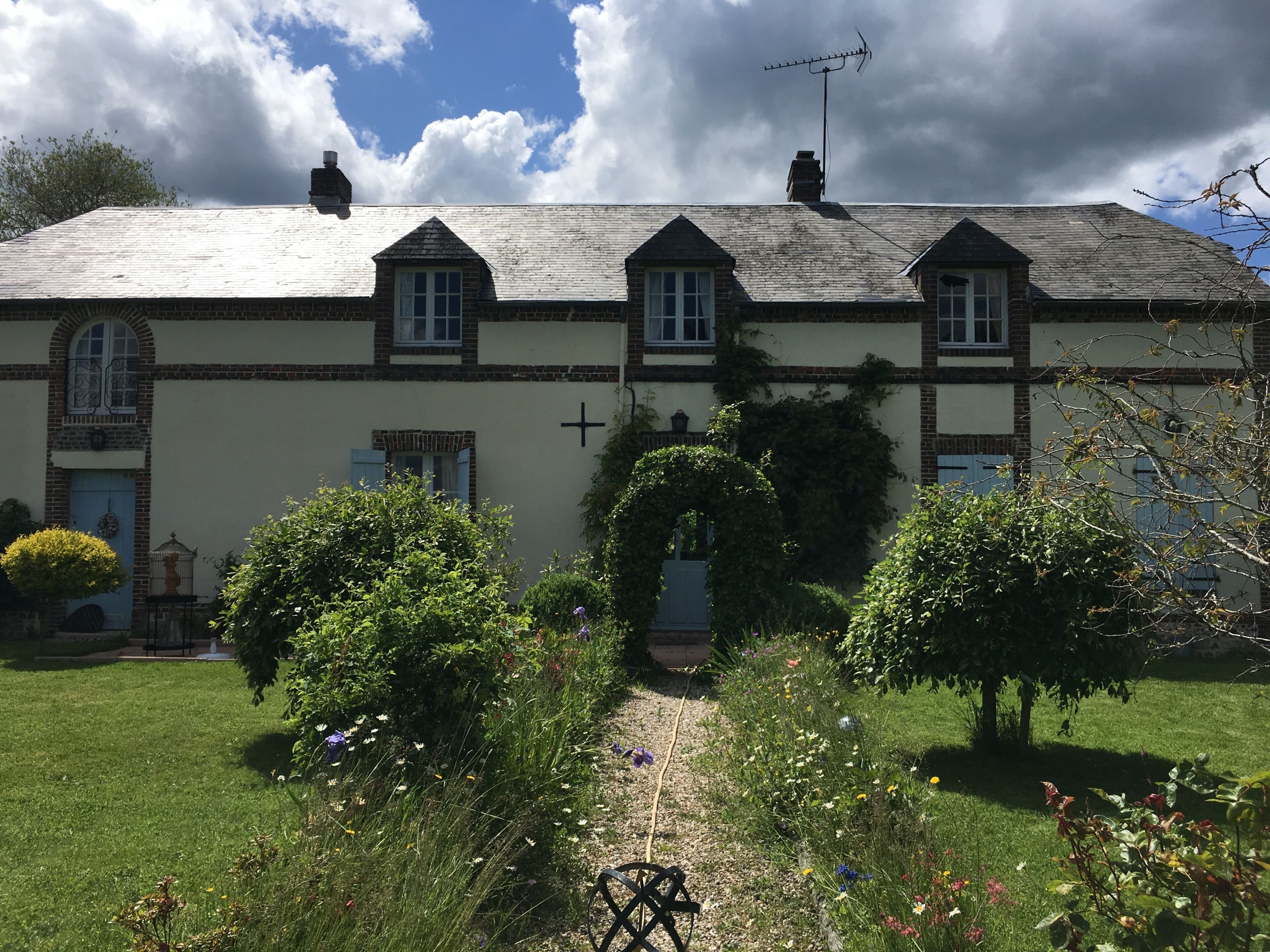 A two-story building with a cross on the wall, a garden with various plants and flowers, and a bicycle in the foreground.
