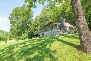 A two-story house with a covered porch and a chimney, surrounded by lush green trees and grass. - 🦌 Wildlife Cove, Wooded Family Cabin with Arcade Game Room, Sleeps 12 (Sevierville)