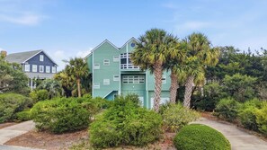 A two-story house with a green exterior, surrounded by lush greenery and palm trees.