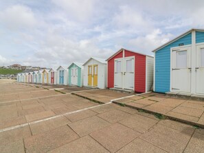 Fileira de cabanas coloridas na praia, com portas e janelas brancas, numeradas em sequência, em área pavimentada.