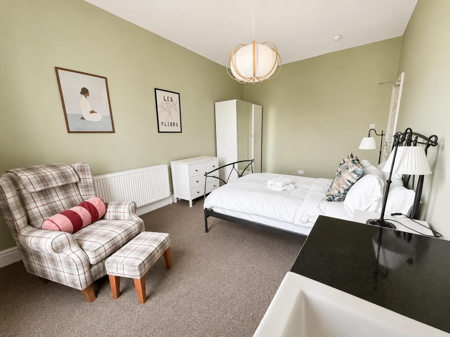 A bedroom with a bed, a chair, a bedside table, a radiator, a wardrobe, and a hanging lamp.