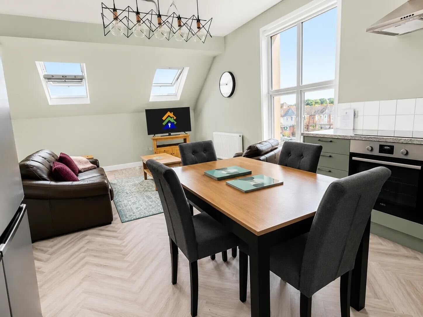 A modern kitchen with a built-in oven, a dining area with a wooden table and chairs, a sofa, and a TV on a stand.