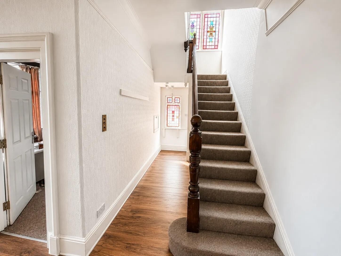A hallway with a carpeted staircase, white walls, and a door leading to a room with a window.