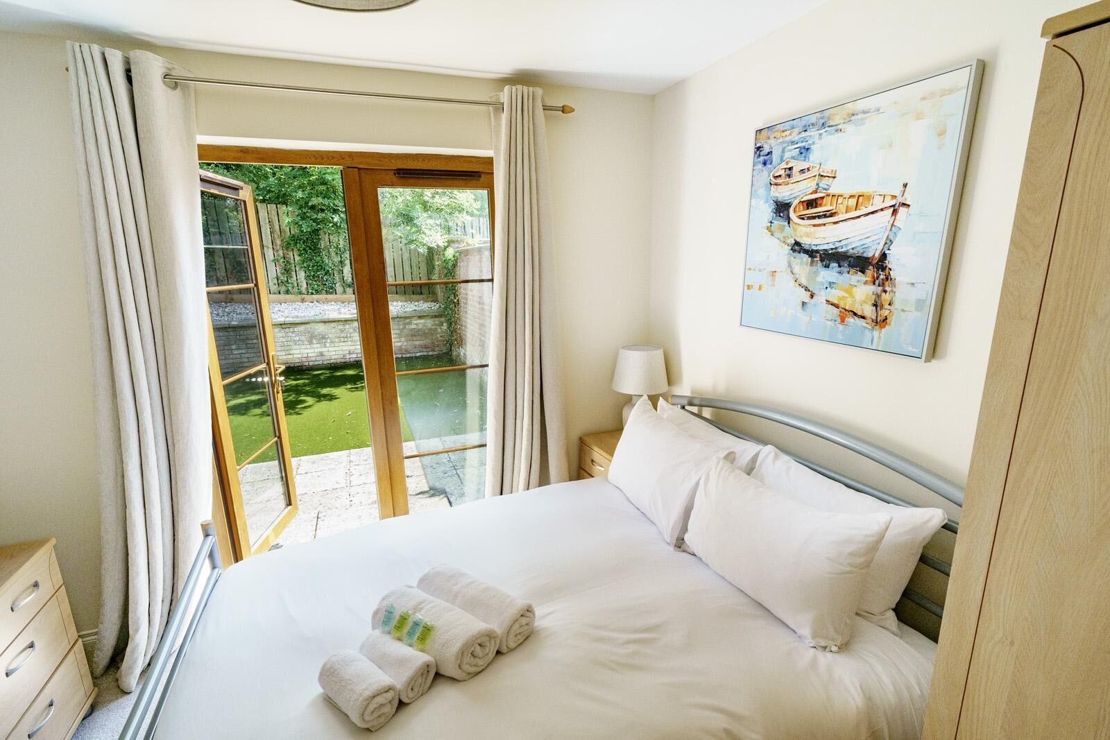 A hotel room with a bed, a painting, a nightstand, a lamp, and a view of a garden through a glass door.