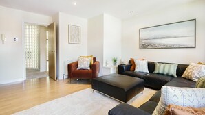 Ein modernes Wohnzimmer mit Sofa, Sesseln und Hocker, einem großen Gemälde an der Wand und einer Tür zu einem weiteren Raum.