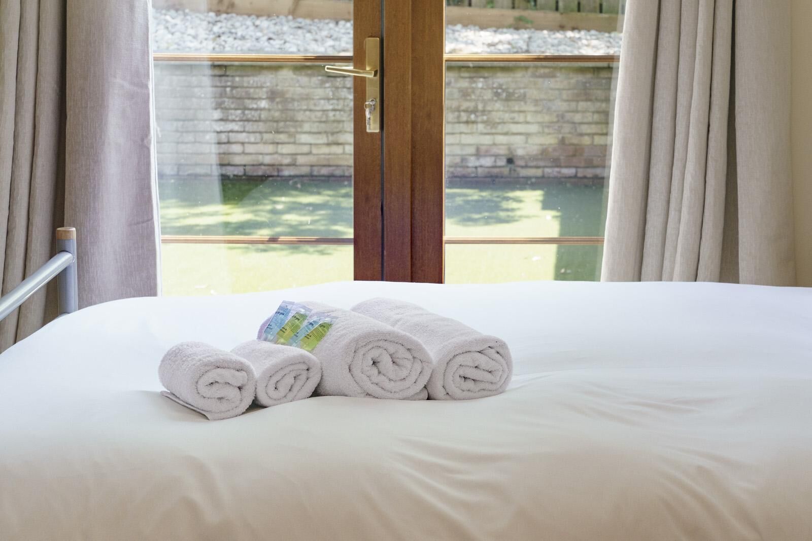 A bed with white linens and rolled towels, a wooden door, and a view of a brick wall and greenery outside.