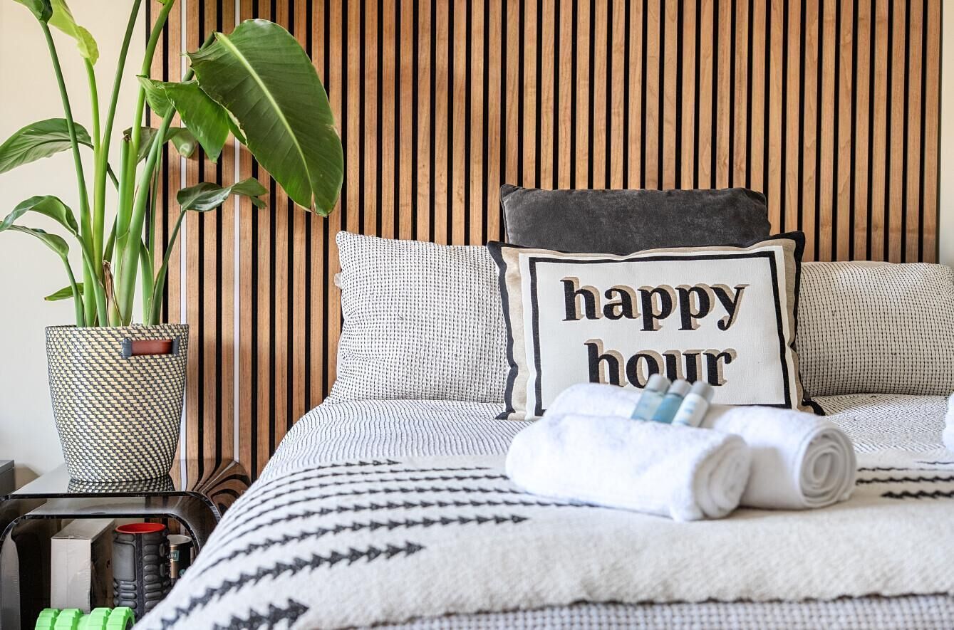 Een netjes opgemaakt bed met een kussen waarop 'happy hour' staat, een potplant en een klein tafeltje met een mok en een vaasje.