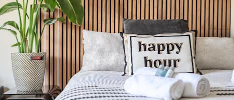 A neatly made bed with a pillow that reads 'happy hour,' a potted plant, and a small table with a mug and a container.