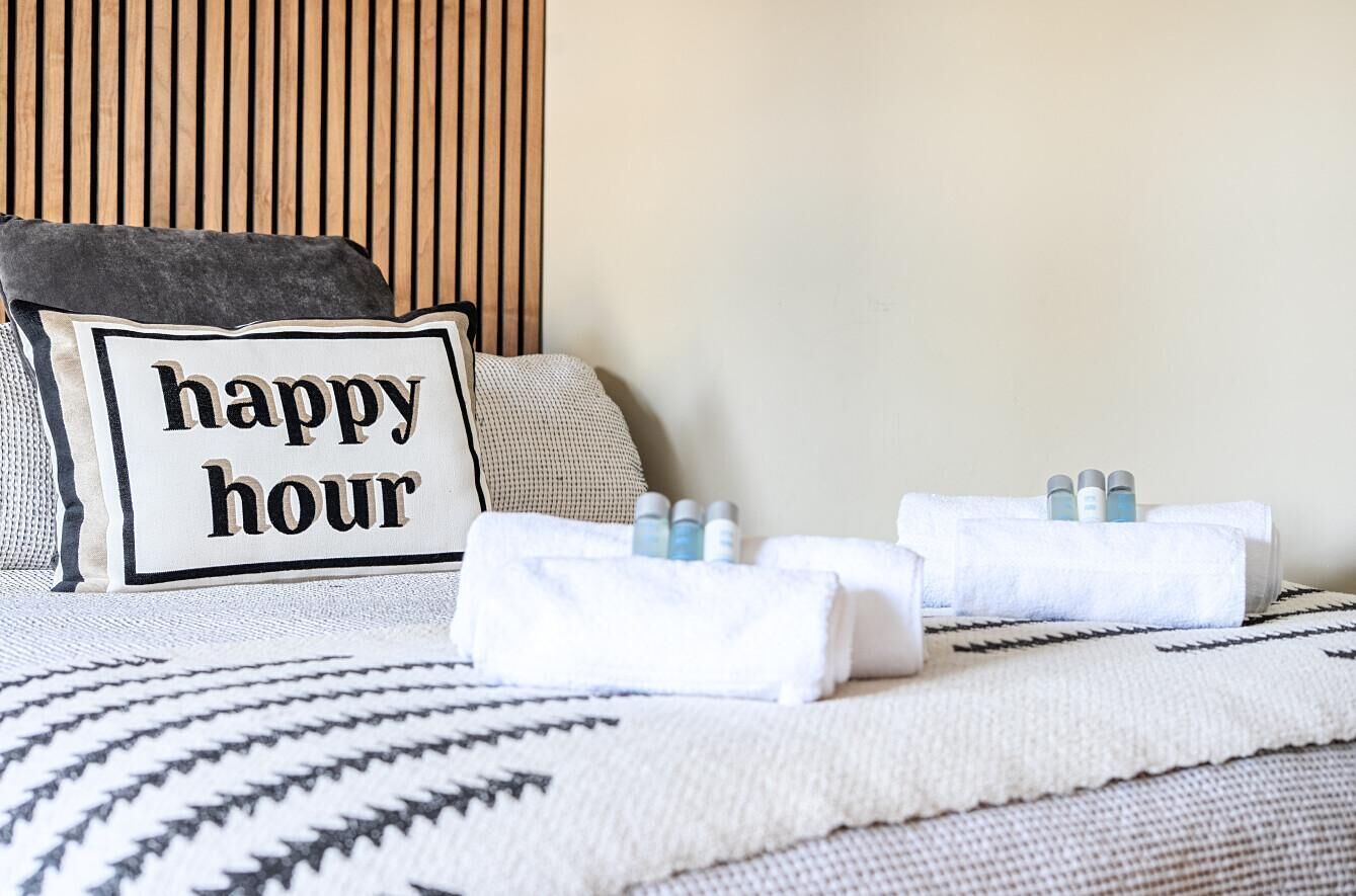 Un lit avec un oreiller orné de l’inscription « happy hour », une serviette et une bouteille de lotion.