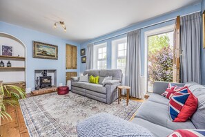 Ein Wohnzimmer mit einer Couch, einem roten Hocker, einem gemusterten Teppich, einem Kamin und einem großen Fenster mit Vorhängen.