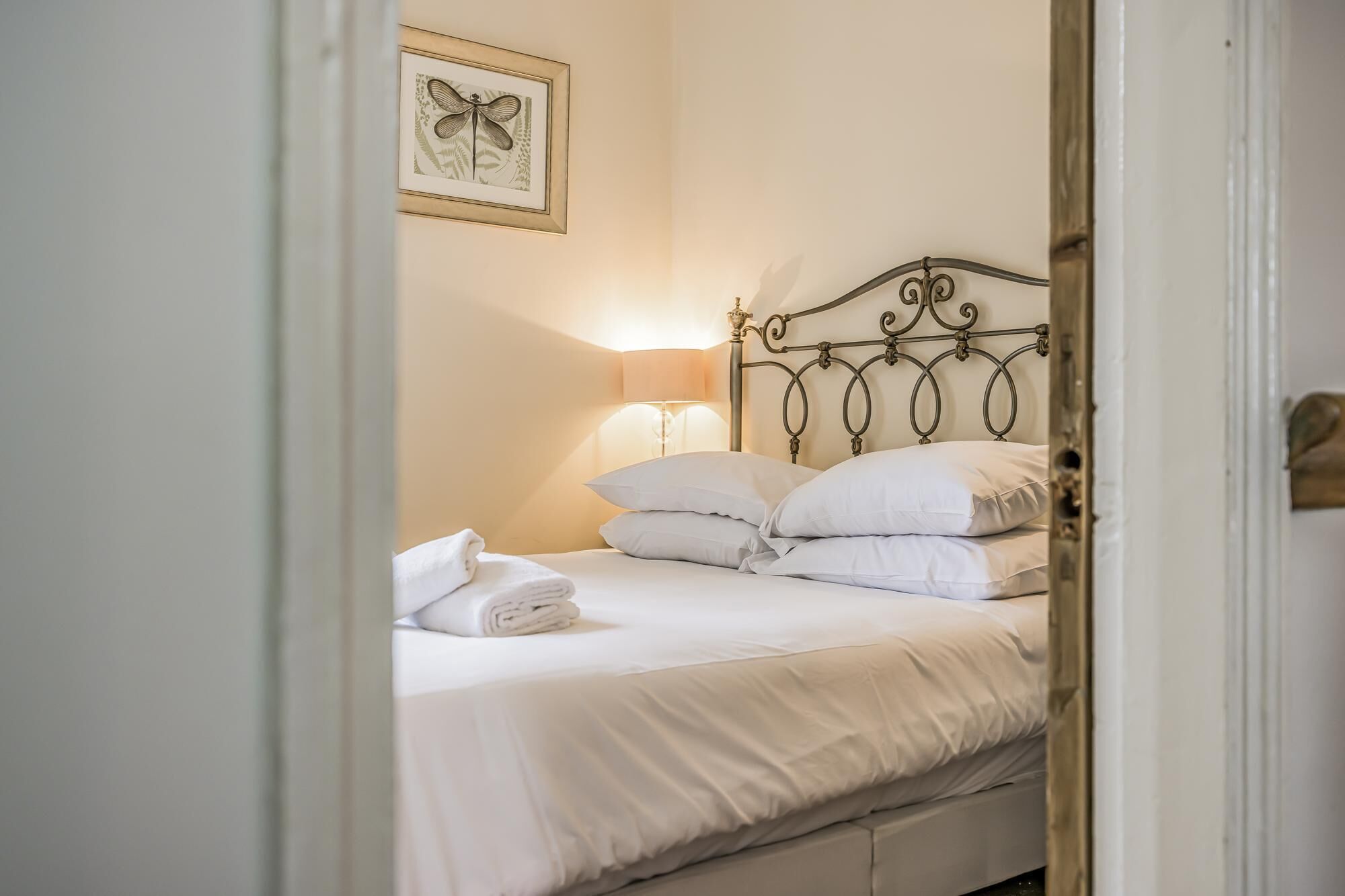 A neatly made bed with white linens, a metal headboard, and a framed botanical print on the wall.
