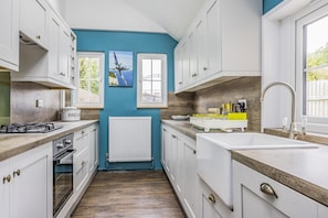 A narrow kitchen with white cabinets, a blue wall, a window, and a radiator. - Number 2 Admiralty Cottages - Pass the Keys (Gosport)