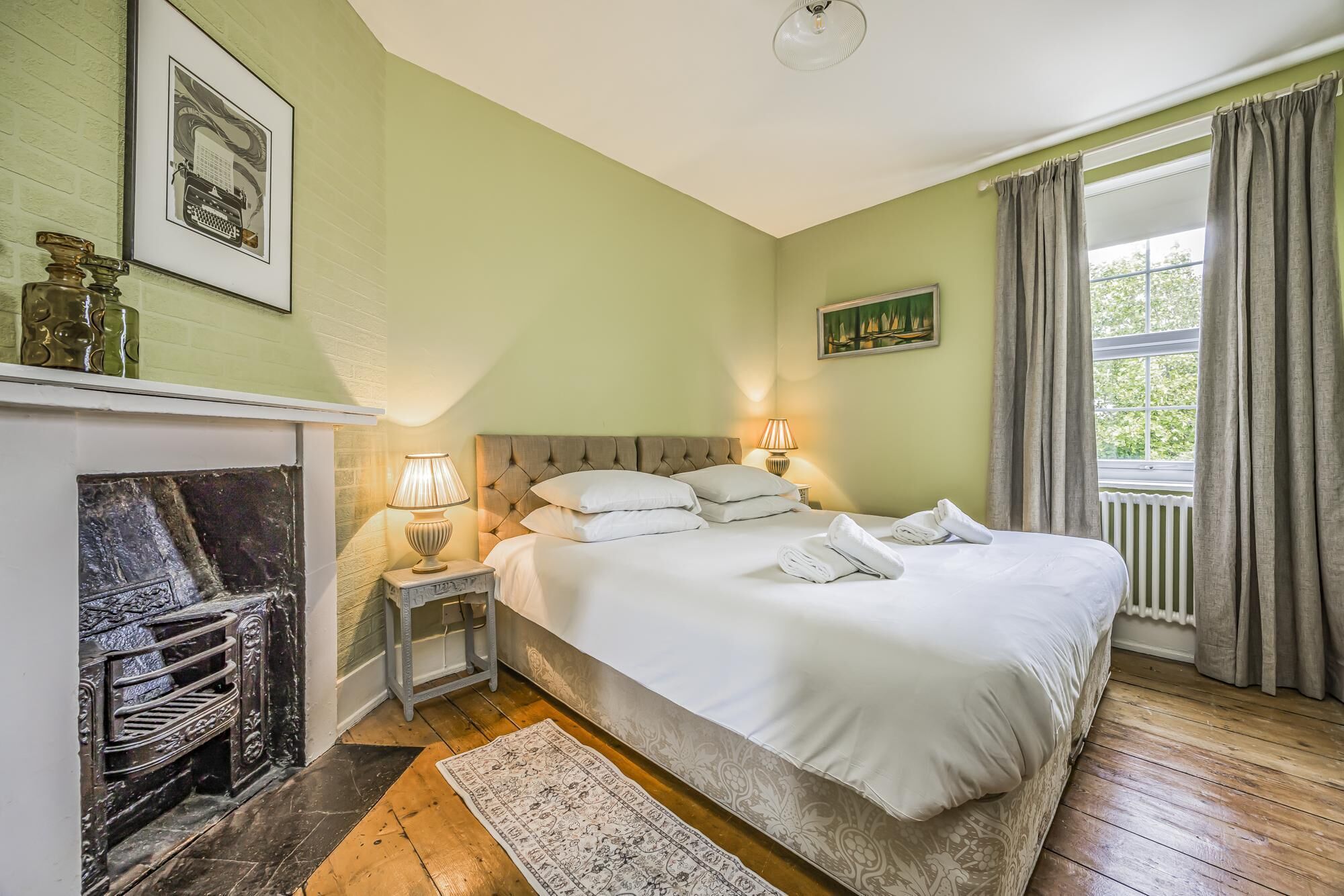 A bedroom with a bed, two bedside lamps, a fireplace, a window with curtains, and a painting on the wall.