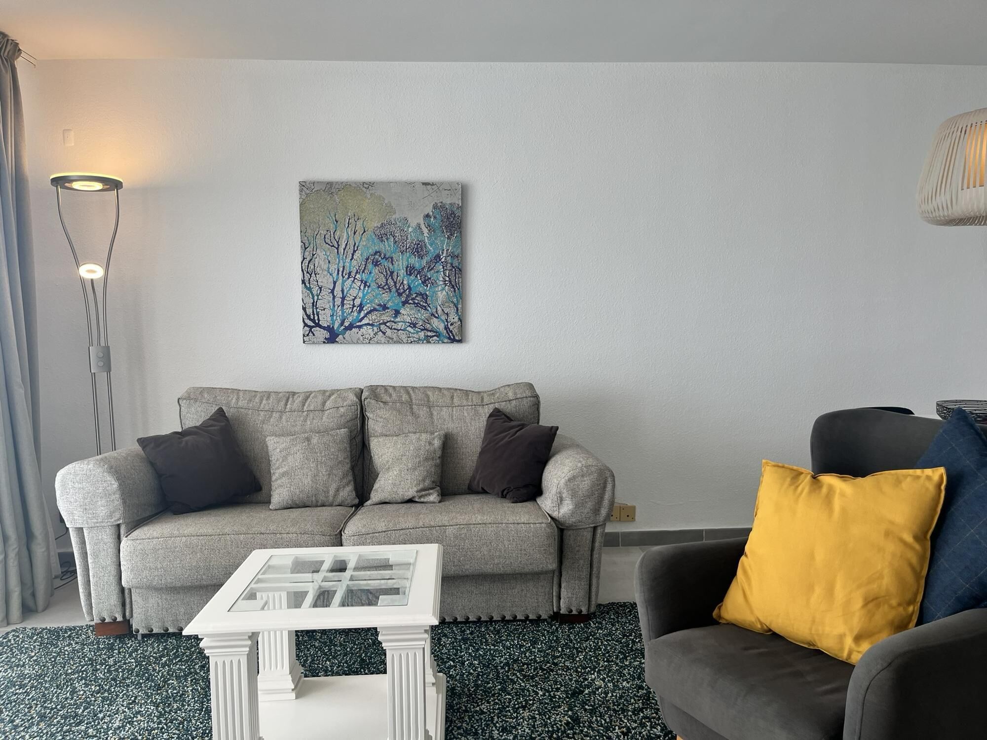 Uma sala de estar com um sofá cinzento, uma mesinha branca, uma poltrona preta com um travesseiro amarelo e um quadro na parede.