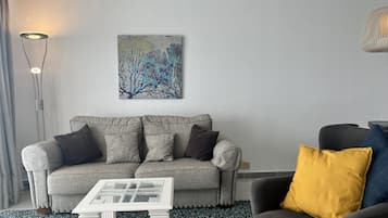 Uma sala de estar com um sofá cinzento, uma mesinha branca, uma poltrona preta com um travesseiro amarelo e um quadro na parede.