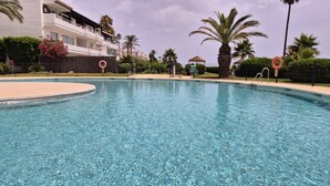 A clear swimming pool with a poolside area, palm trees, and a building with balconies. - Beachfront Escape in Miraflores - Pass the Keys (Las Lagunas de Mijas)