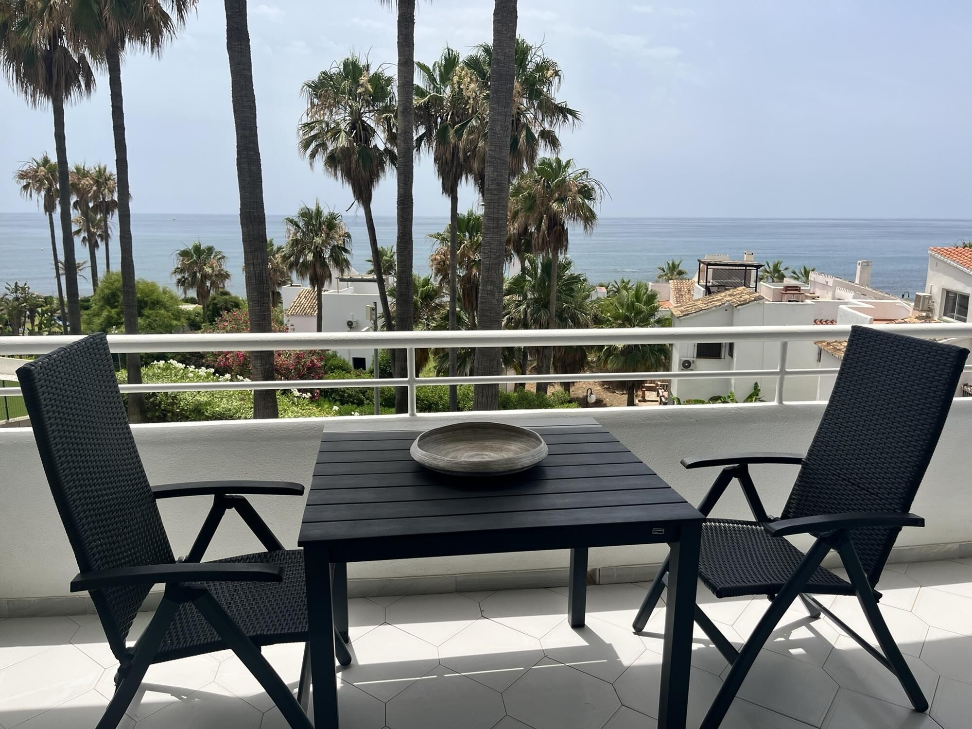 Un balcón con dos sillas y una mesa, con vista al paisaje costero que incluye palmeras y edificios.
