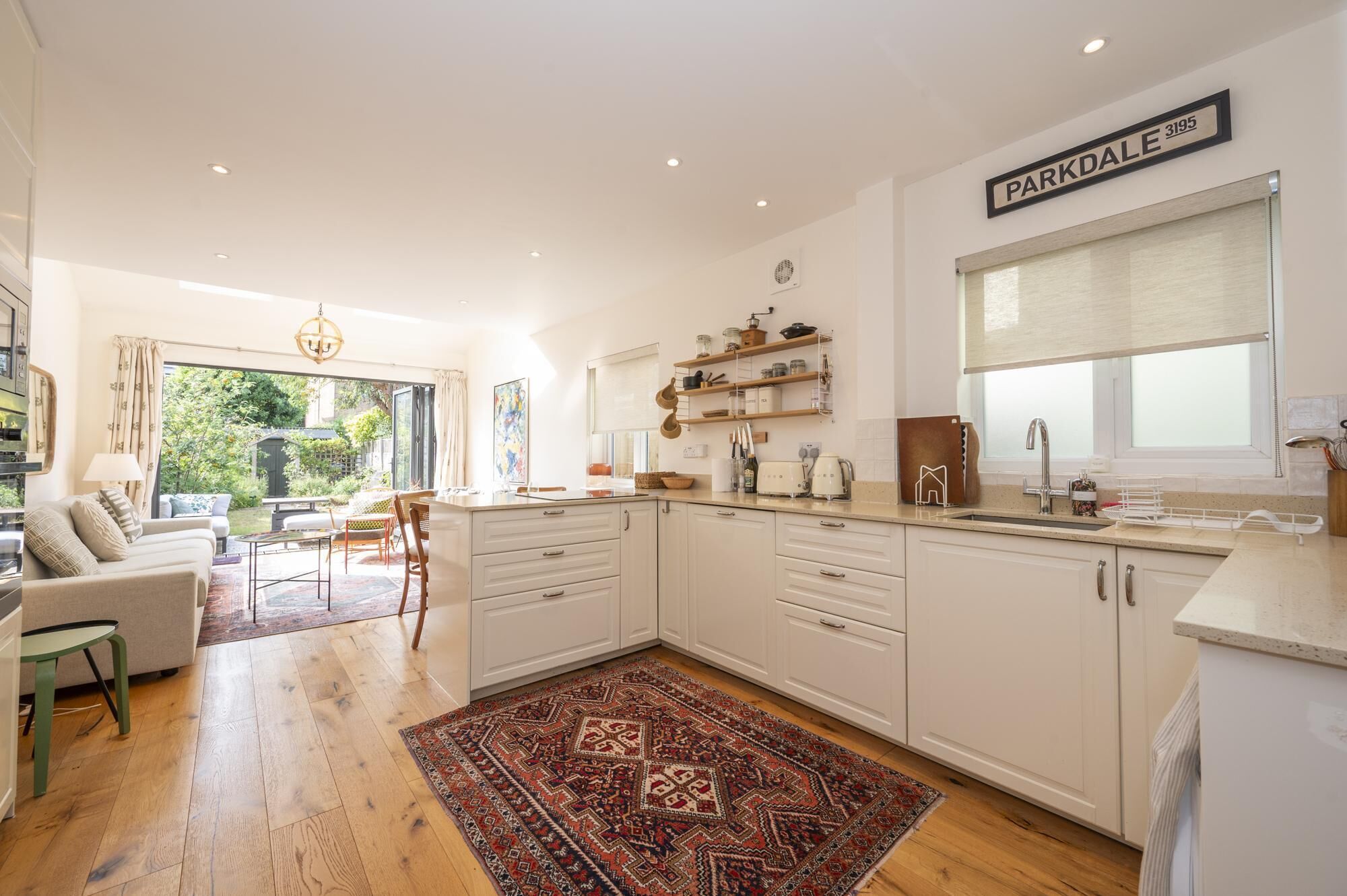 Una cucina moderna con mobili bianchi, un'area pranzo con divano e poltrone, e vista su un giardino attraverso ampie finestre.