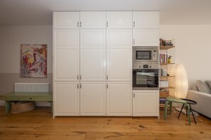 Una habitación con un armario blanco, un radiador, un banco verde, una mesa, una lámpara, un sofá y una estantería con libros y objetos decorativos.