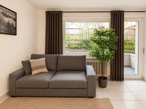 Un sofá gris con un cojín, una planta en maceta y una ventana con cortinas.