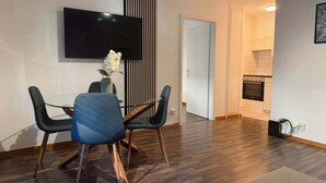 A modern living space with a dining area featuring a glass table and chairs, a flat-screen TV mounted on the wall, and a kitchen with a built-in oven. - book2stay: Apartment 2.5-room Biel (Biel)