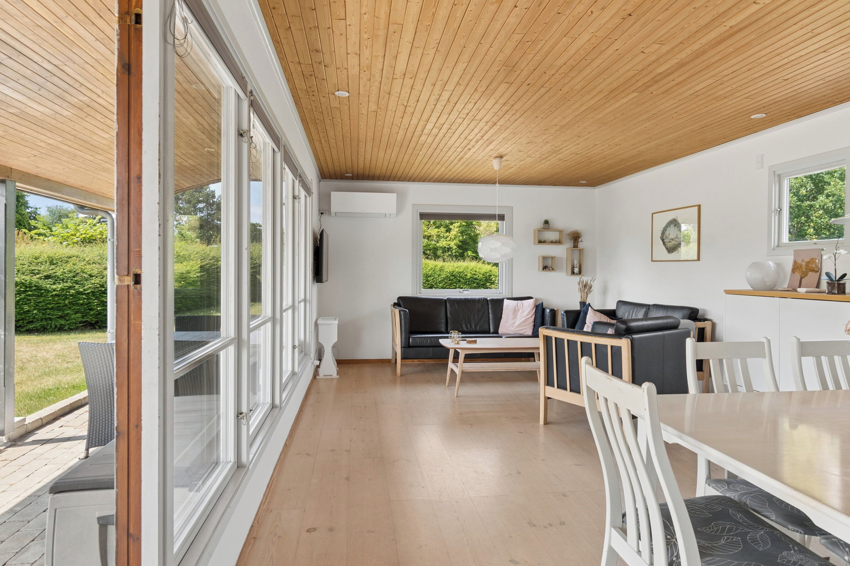 A spacious room with a wooden ceiling, large windows, a black leather sofa, a dining table with chairs, and a white wall with a picture.