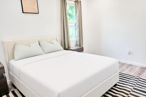 Ein ordentlich bezogenes Bett in einem Zimmer mit Fenster, Nachttisch und Teppich in Schwarz-Weiß-Streifen.