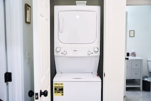 Una lavadora y secadora blancas con panel de control, puerta y un pequeño armario con un cubo de basura.
