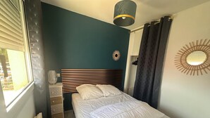 Et soveværelse med seng, et natbord, en lampe, et vindue med persienner og en dekorativ genstand på væggen.