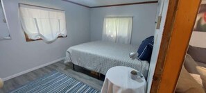 A bedroom with a bed, a bedside table, a window with curtains, a small round table, and a rug. - West Battle Seasonal Cabin (Henning)