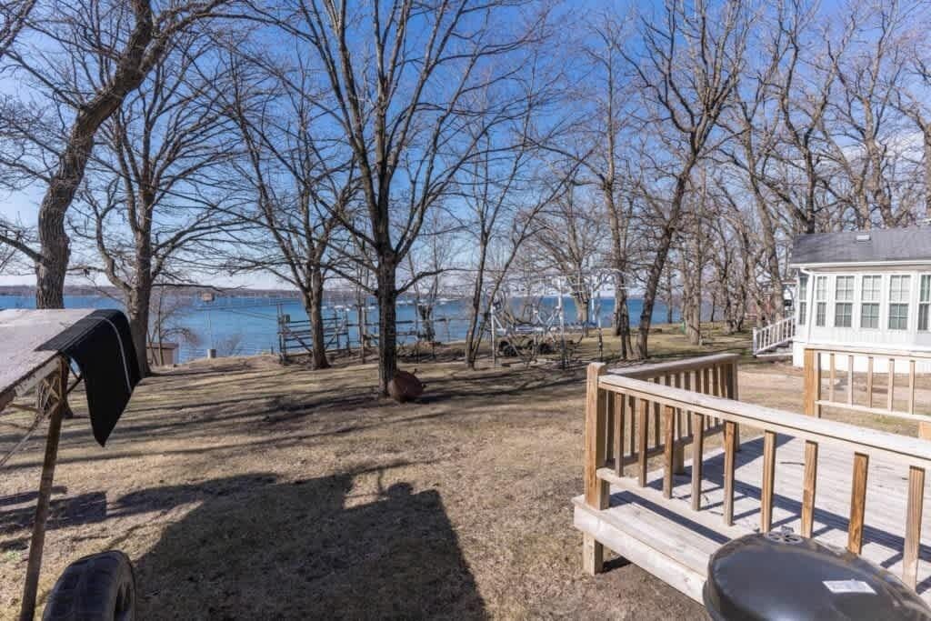 A wooden deck with a grill, a white building with windows, and leafless trees by a body of water.