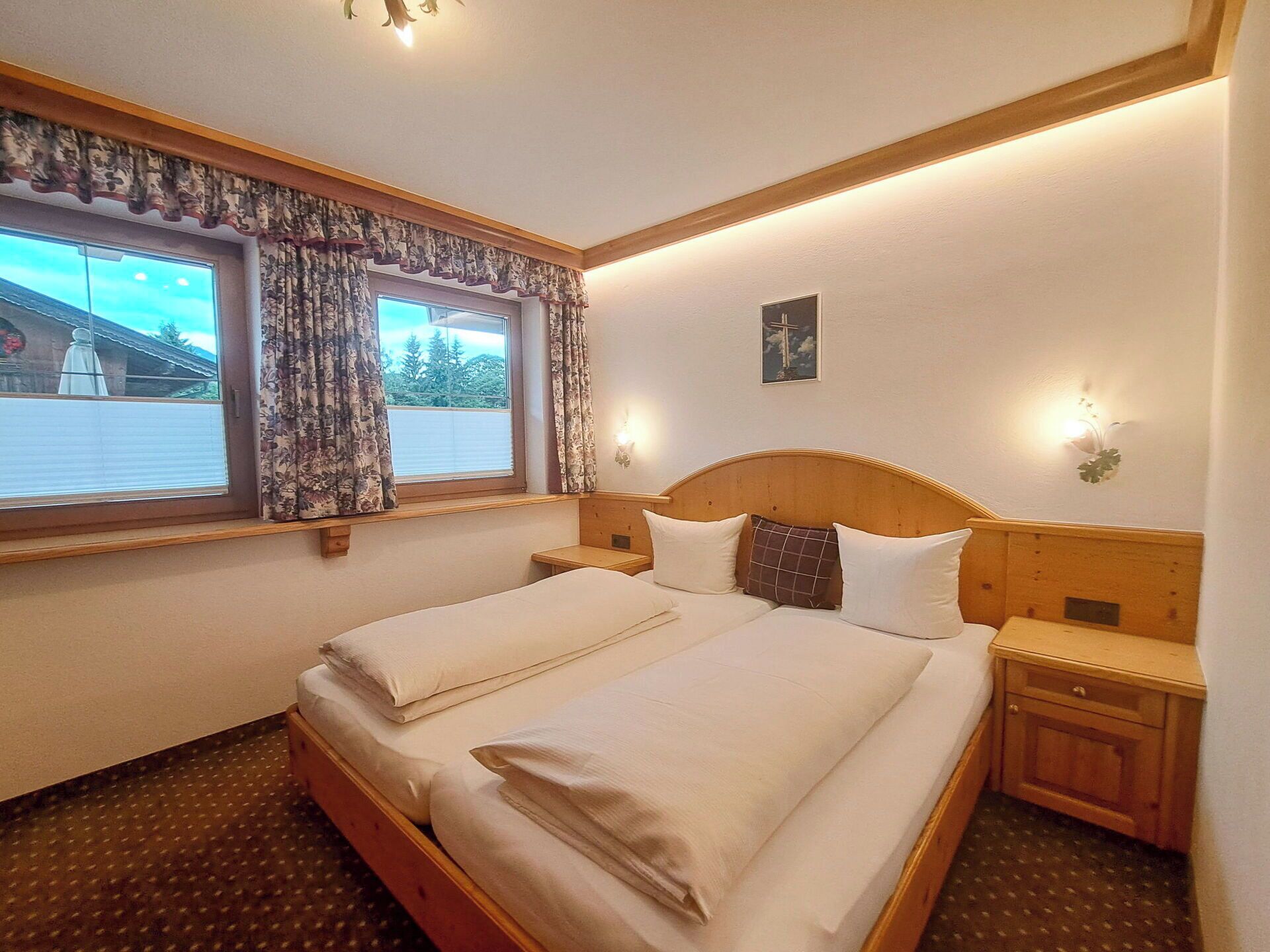 A double bed with headboards, a window with curtains, a wall-mounted light, and a small bedside table.