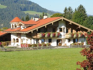 Ein traditionelles Chalet mit rotem Ziegeldach, mehreren Balkonen mit Blumenkästen und Blick auf eine Berglandschaft.