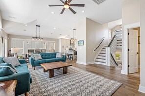 A spacious living area with a sofa set, coffee table, patterned rug, ceiling fan, and a staircase leading to an upper level. - Steps to Sand-Full Remodel Like New! (Corolla)