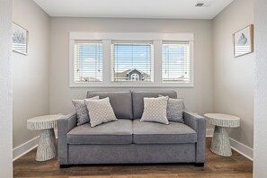 A grey sofa with patterned cushions, two round side tables, and a window with blinds overlooking a building. - Steps to Sand-Full Remodel Like New! (Corolla)
