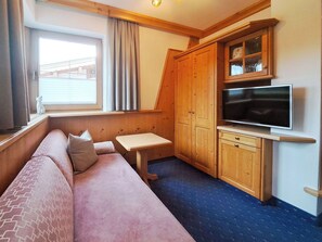 Ein gemütliches Zimmer mit einer Couch, einem kleinen Tisch, einem Fernseher, Holzschränken und einem Fenster mit Vorhängen.