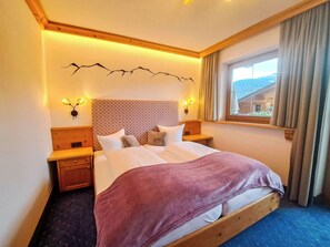 Quarto de hotel com cama, duas mesas de cabeceira, cabeceira com padrão decorativo, luminárias fixadas na parede, janela com cortinas e vista para um edifício.