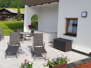Um pátio externo com móveis de vime, uma mesa e vasos com flores.