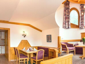 Ein gemütliches Zimmer mit Holztisch, violetten Polsterstühlen, einer Holzbank und einem Fenster mit blumigen Vorhängen.
