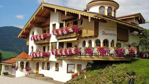 Un edificio de varios pisos con balcones decorados con flores, un cartel que dice 'Appartments Alpenschlöfi' y un paisaje montañoso de fondo.